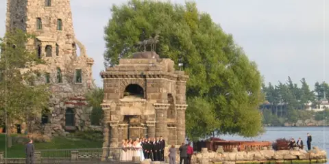 A wedding party poses in front of the stone Dove-Cote structure with the main tower of Boldt Castle visible in the background.