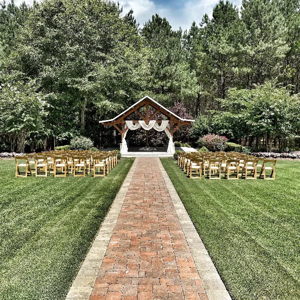 Outdoor wedding ceremony setup with a wooden arbor draped in white fabric and rows of chairs on a grassy lawn surrounded by trees.