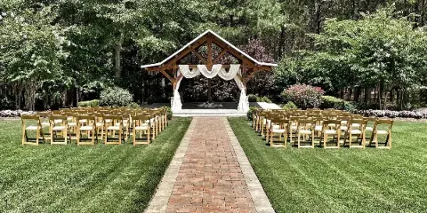 Outdoor wedding ceremony setup with a wooden arbor draped in white fabric and rows of chairs on a grassy lawn surrounded by trees.