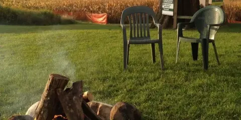 A campfire pit with logs and chairs on a grassy lawn, with a corn field and a maze rules sign in the background.