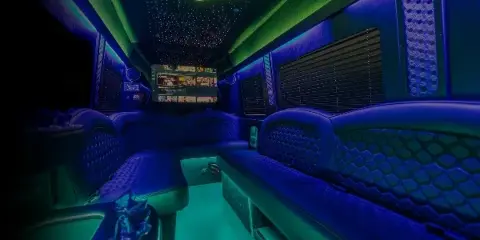 Interior view of a party bus with blue and green ambient lighting, quilted leather seating, and a mounted television screen.