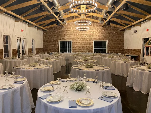 Interior view of a rustic wedding venue featuring exposed wooden ceiling beams, brick walls, and round tables set with white linens and floral centerpieces.