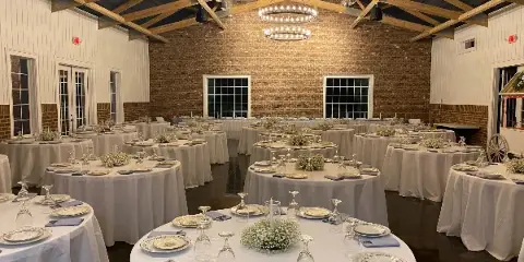 Interior view of a rustic wedding venue featuring exposed wooden ceiling beams, brick walls, and round tables set with white linens and floral centerpieces.