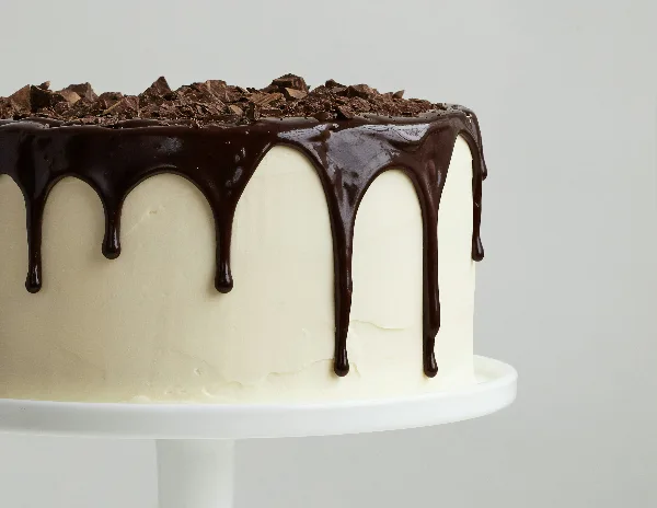 A close-up photograph of a white frosted cake with a chocolate drip and chocolate shavings on top, sitting on a white cake stand.
