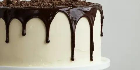 A close-up photograph of a white frosted cake with a chocolate drip and chocolate shavings on top, sitting on a white cake stand.