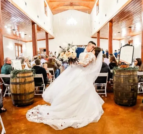 Bride and groom kissing during a wedding ceremony inside a rustic barn with guests seated in the background.
