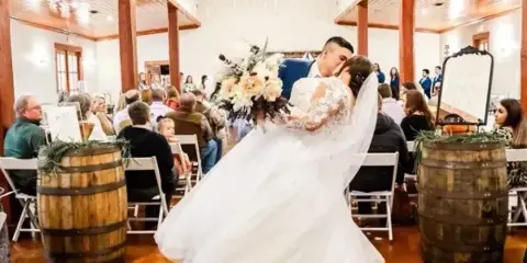 Bride and groom kissing during a wedding ceremony inside a rustic barn with guests seated in the background.