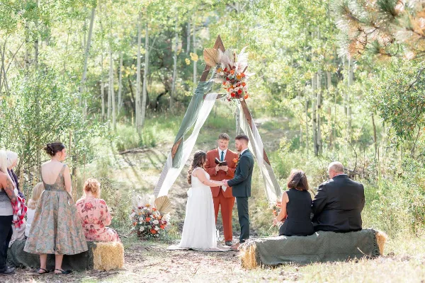 A bride and groom exchanging vows under a floral arch in a forest setting with seated guests.