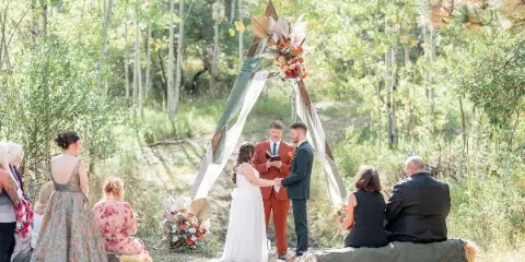 A bride and groom exchanging vows under a floral arch in a forest setting with seated guests.