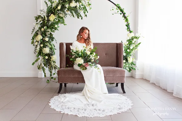 Bride seated on a brown lounge bench beneath a floral arch with white flowers and greenery in a bright indoor venue.