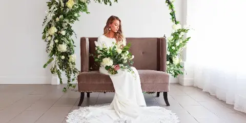 Bride seated on a brown lounge bench beneath a floral arch with white flowers and greenery in a bright indoor venue.