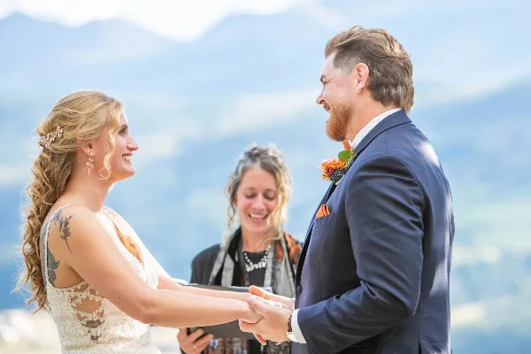 A bride and groom holding hands during a wedding ceremony with an officiant standing between them, set against a mountain backdrop.