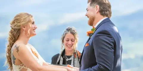 A bride and groom holding hands during a wedding ceremony with an officiant standing between them, set against a mountain backdrop.