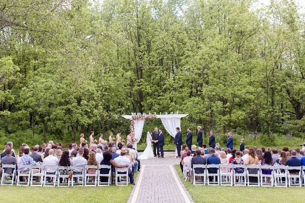 Outdoor wedding ceremony with bride and groom at a floral altar surrounded by seated guests and a wooded background.