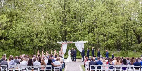 Outdoor wedding ceremony with bride and groom at a floral altar surrounded by seated guests and a wooded background.