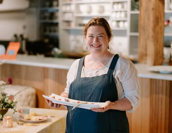 A smiling woman in a dark apron holds a white platter of plated food in a kitchen setting.