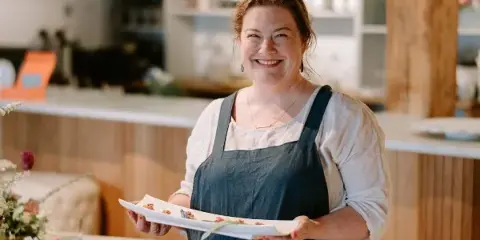 A smiling woman in a dark apron holds a white platter of plated food in a kitchen setting.