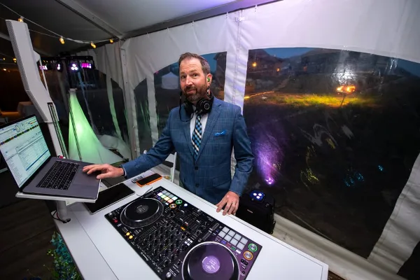 A professional wedding DJ in a blue suit standing behind a Pioneer DJ setup and laptop at an outdoor event.