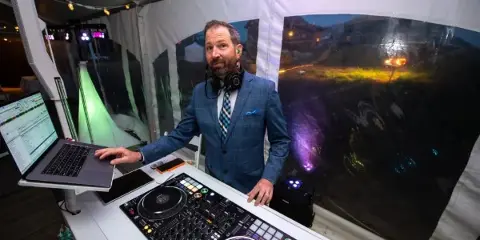 A professional wedding DJ in a blue suit standing behind a Pioneer DJ setup and laptop at an outdoor event.