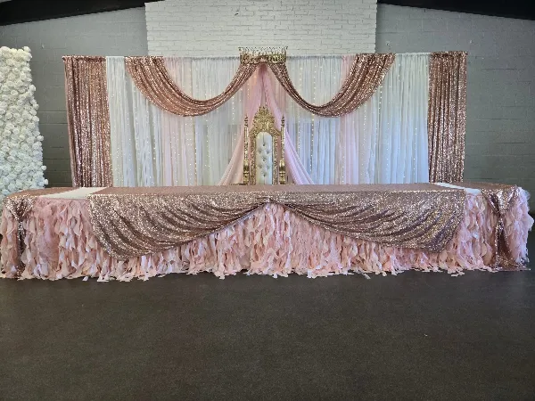 A decorated event head table featuring a gold throne chair, rose gold drapery, and a white floral wall.