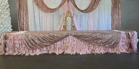 A decorated event head table featuring a gold throne chair, rose gold drapery, and a white floral wall.