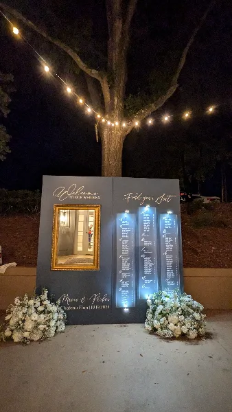 A wedding event setup at night featuring a large grey backdrop with a gold-framed mirror, a 'Welcome to your wedding' sign, and a seating chart, decorated with white flowers and string lights.