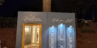 A wedding event setup at night featuring a large grey backdrop with a gold-framed mirror, a 'Welcome to your wedding' sign, and a seating chart, decorated with white flowers and string lights.