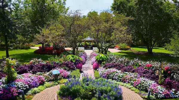 A lush park garden featuring a central brick pathway, vibrant flower beds, and a wooden gazebo in the background.