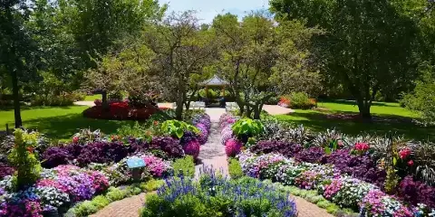 A lush park garden featuring a central brick pathway, vibrant flower beds, and a wooden gazebo in the background.