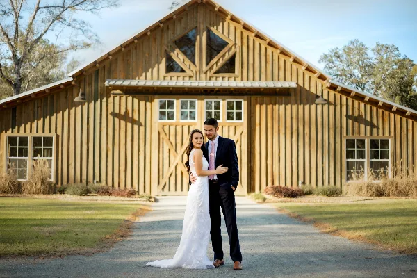 A bride and groom standing in front of a large rustic wooden barn with a gravel driveway and grassy surroundings.
