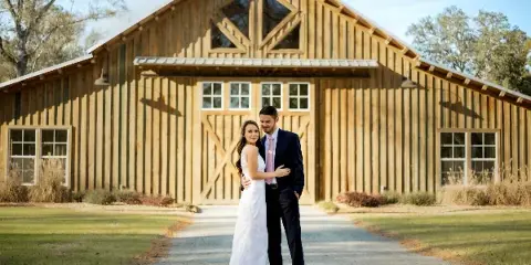 A bride and groom standing in front of a large rustic wooden barn with a gravel driveway and grassy surroundings.