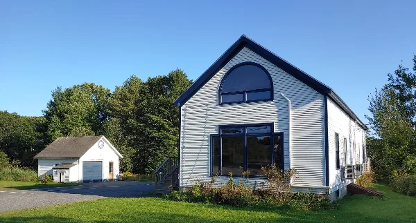 Exterior view of a white community center building with a large arched window and a smaller garage-like structure, set on a grassy lawn.