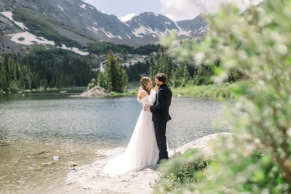 A bride and groom standing together by a mountain lake in a wedding dress and suit.