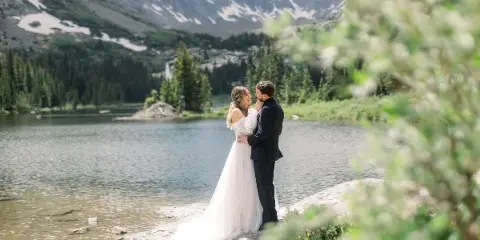 A bride and groom standing together by a mountain lake in a wedding dress and suit.