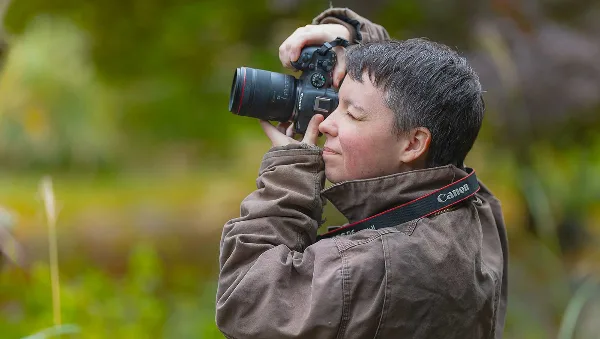 A photographer with short hair wearing a brown jacket holds a Canon DSLR camera up to their eye, taking a photo in a natural outdoor environment.