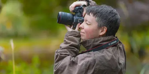 A photographer with short hair wearing a brown jacket holds a Canon DSLR camera up to their eye, taking a photo in a natural outdoor environment.