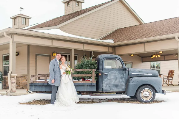 A bride and groom posing in front of a rustic barn venue with a vintage truck in the snow.