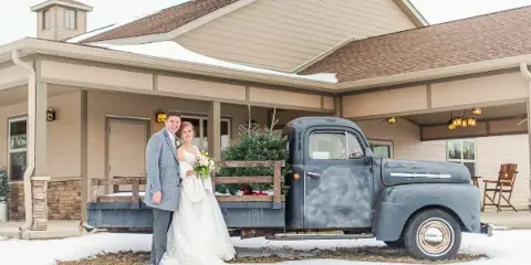 A bride and groom posing in front of a rustic barn venue with a vintage truck in the snow.