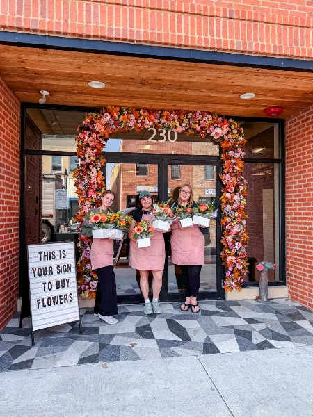 Three staff members in pink aprons holding flower bouquets standing in front of a brick storefront decorated with a large floral arch.