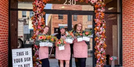 Three staff members in pink aprons holding flower bouquets standing in front of a brick storefront decorated with a large floral arch.