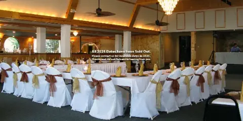 Interior view of a wedding reception hall with long tables, white chair covers, and wooden beams.