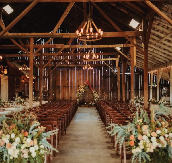 Interior of a rustic barn wedding venue featuring exposed wooden beams, hanging chandeliers, and rows of chairs set up for a ceremony.