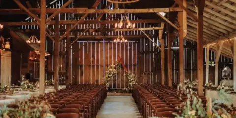 Interior of a rustic barn wedding venue featuring exposed wooden beams, hanging chandeliers, and rows of chairs set up for a ceremony.