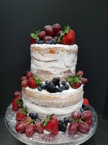 Two-tier semi-naked cake with white frosting, topped and surrounded by fresh strawberries, red grapes, blueberries, and blackberries on a silver cake board against a dark background.