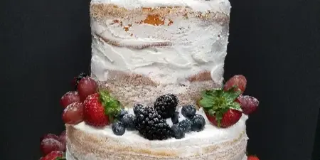 Two-tier semi-naked cake with white frosting, topped and surrounded by fresh strawberries, red grapes, blueberries, and blackberries on a silver cake board against a dark background.