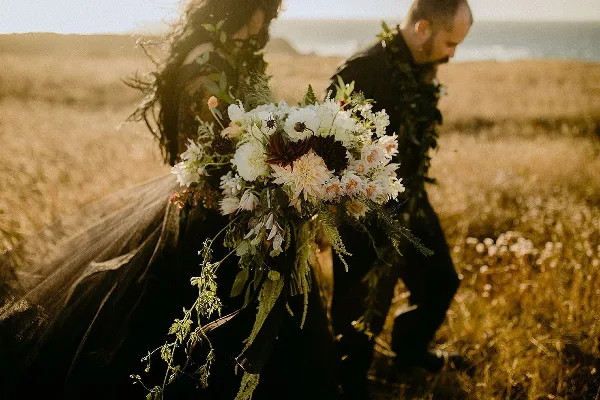A bride and groom in a coastal field holding a large bouquet of white and dark dahlias, chrysanthemums, and greenery.