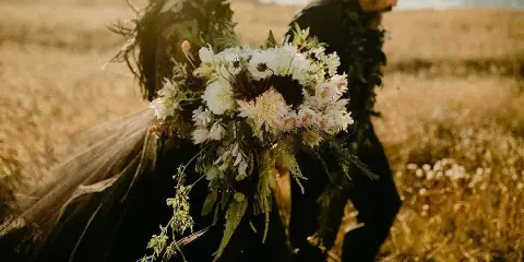 A bride and groom in a coastal field holding a large bouquet of white and dark dahlias, chrysanthemums, and greenery.