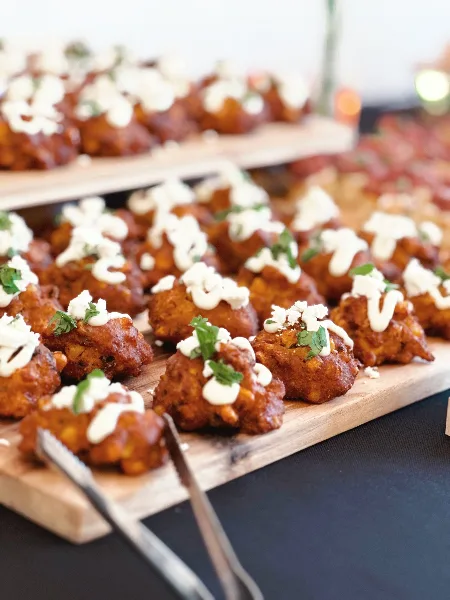 A close-up of a catering dish featuring fried bites topped with white sauce and herbs, served on a wooden board.