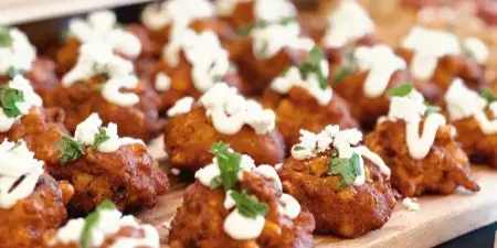 A close-up of a catering dish featuring fried bites topped with white sauce and herbs, served on a wooden board.