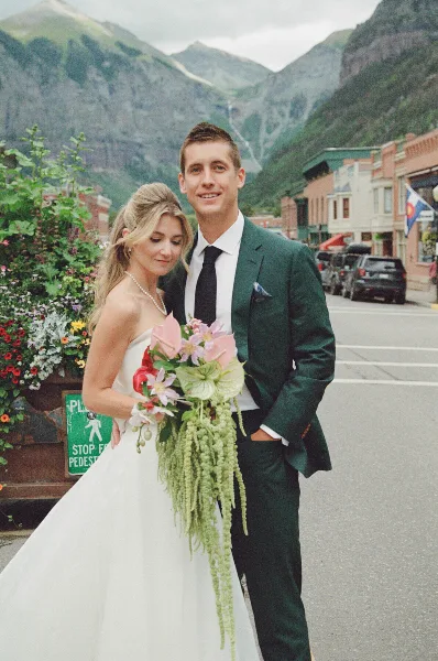 A bride and groom posing on a street in a mountain town with a bouquet and green suit.